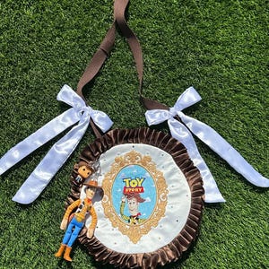 May include: A round brown and white bag featuring a Toy Story graphic with Woody inside an ornate gold frame. The bag has a ruffled brown border, two white satin bows, a brown shoulder strap, and a small Woody figurine attached to the side.