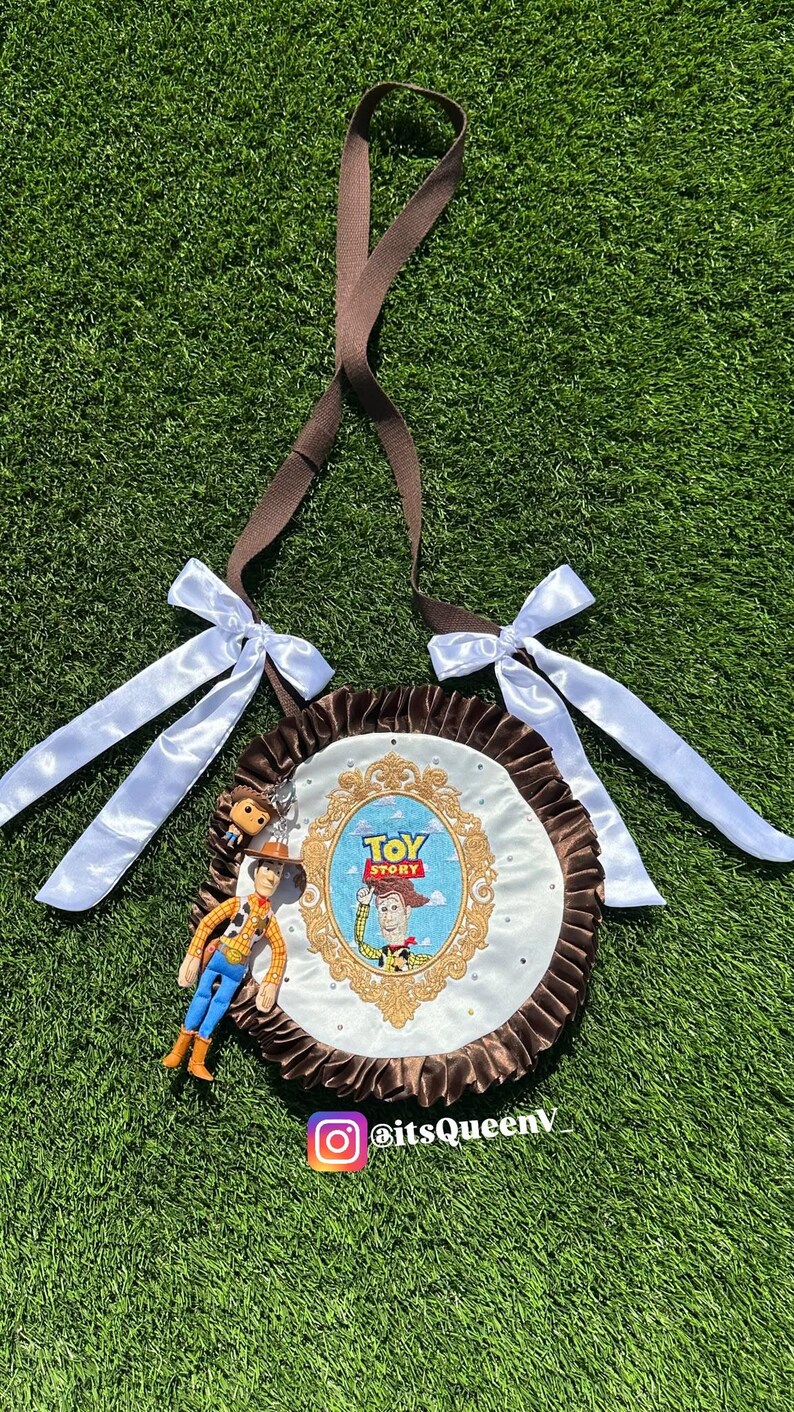 May include: A round brown and white bag featuring a Toy Story graphic with Woody inside an ornate gold frame. The bag has a ruffled brown border, two white satin bows, a brown shoulder strap, and a small Woody figurine attached to the side.