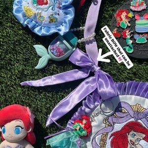 Op de afbeelding: Ariel-thema accessoires, waaronder een pluche pop, een blauwe satijnen strik en een decoratieve tas met een geborduurd Ariel-portret. De items zijn gerangschikt op een groen oppervlak, met een pluche vis en een verzameling pins.