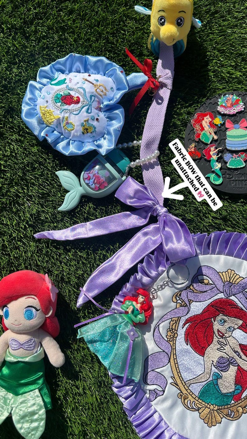 Op de afbeelding: Ariel-thema accessoires, waaronder een pluche pop, een blauwe satijnen strik en een decoratieve tas met een geborduurd Ariel-portret. De items zijn gerangschikt op een groen oppervlak, met een pluche vis en een verzameling pins.