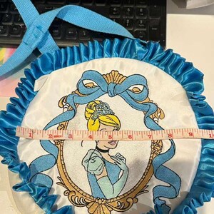 Op de afbeelding: Een ronde, witte tas met een Cinderella-thema met een blauwe satijnen ruchesrand en een blauwe handgreep. De tas heeft een geborduurde afbeelding van Cinderella in een gouden lijst. De tas heeft een diameter van ongeveer 25 cm.
