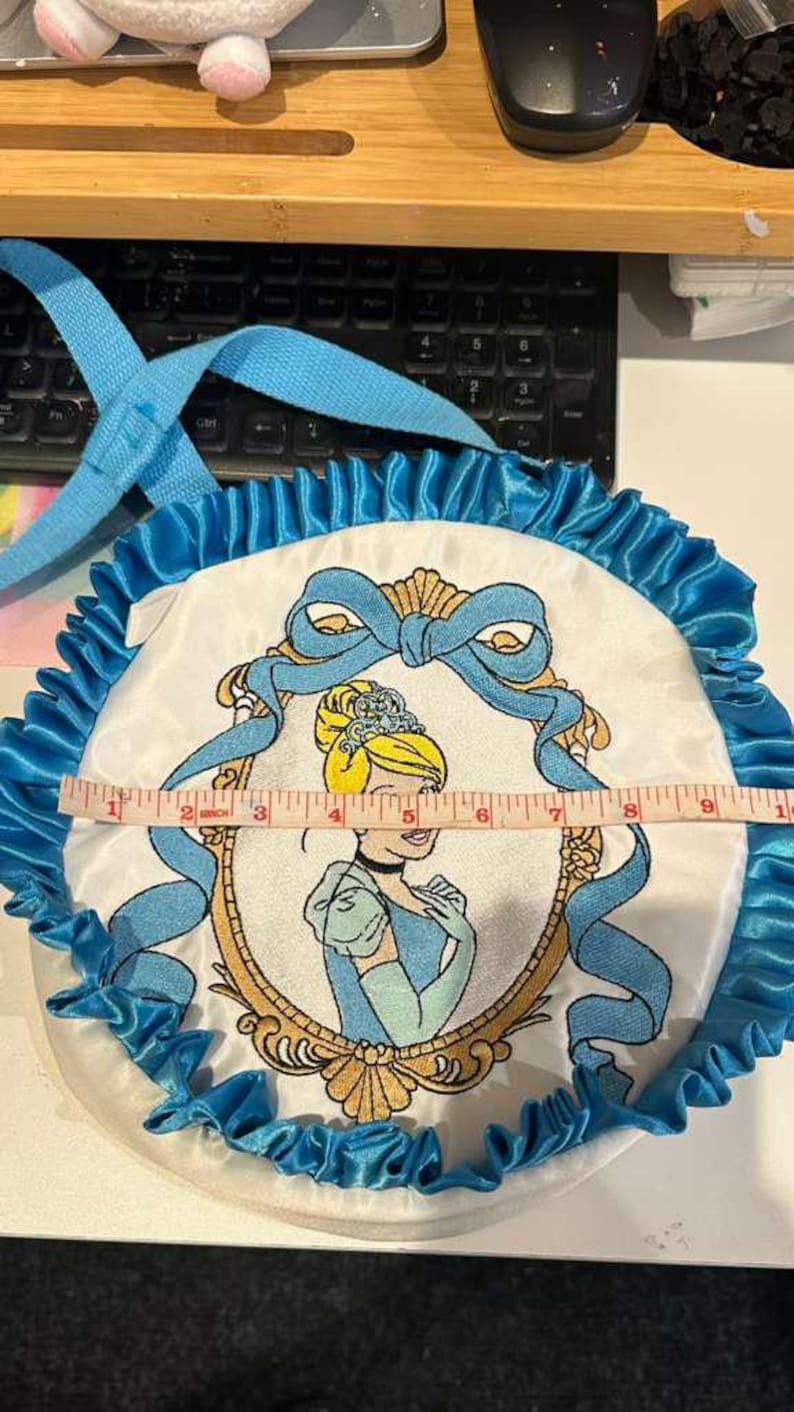 Op de afbeelding: Een ronde, witte tas met een Cinderella-thema met een blauwe satijnen ruchesrand en een blauwe handgreep. De tas heeft een geborduurde afbeelding van Cinderella in een gouden lijst. De tas heeft een diameter van ongeveer 25 cm.