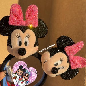 May include: Two Minnie Mouse ear headbands. The ears are black with pink bows. The Minnie Mouse face is plush with embroidered features. The headband is black and blue. The image also includes a small heart-shaped card with a cartoon illustration.