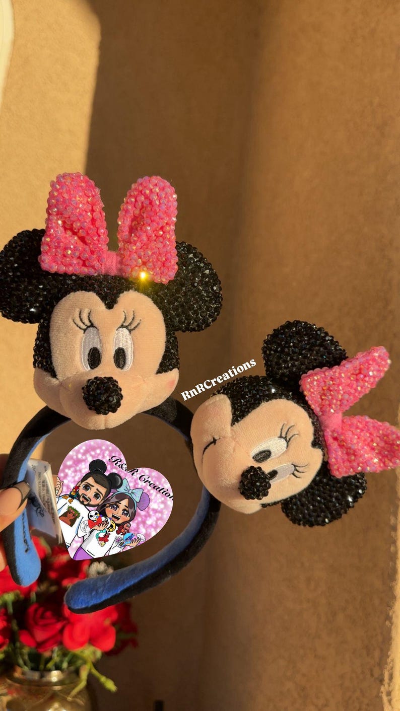 May include: Two Minnie Mouse ear headbands. The ears are black with pink bows. The Minnie Mouse face is plush with embroidered features. The headband is black and blue. The image also includes a small heart-shaped card with a cartoon illustration.