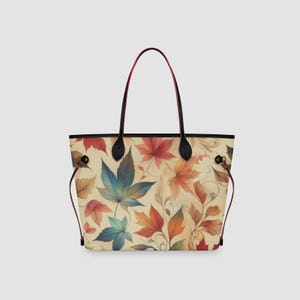 May include: A tote bag featuring a cream-colored background with a repeating pattern of autumn leaves in shades of red, orange, yellow, and blue. The bag has two black shoulder straps with red edges and thin black side cords with red accents.