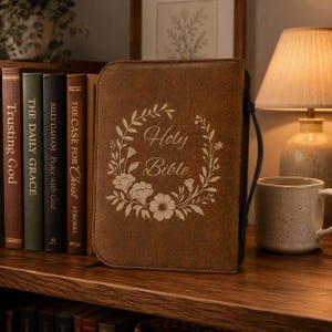 May include: A brown leather Bible cover with the words "Holy Bible" in a floral wreath design. The cover is next to a row of books with titles like "Trusting God" and "The Daily Grace". A mug and lamp are in the background.