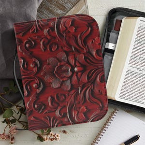Può includere: Una copertina rossa per Bibbia con un motivo floreale in rilievo. La copertina è aperta, rivelando un libro con testo. Sono visibili anche un taccuino e una penna.