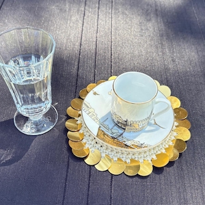May include: A white coffee cup and saucer with a gold rim and a gold and white patterned coaster. The coaster is made of a circle of gold coins.