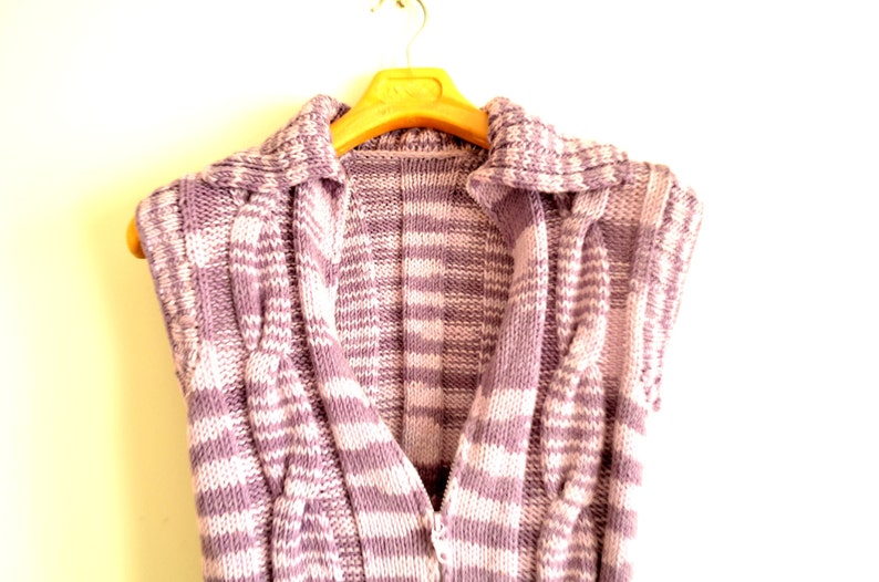 May include: A purple and white striped knitted vest with a collar and a V-neck. The vest is hanging on a wooden hanger.