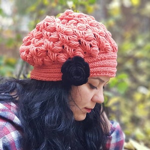 May include: A coral pink crocheted beanie with a black flower embellishment. The beanie is made with a textured stitch and has a slouchy fit.