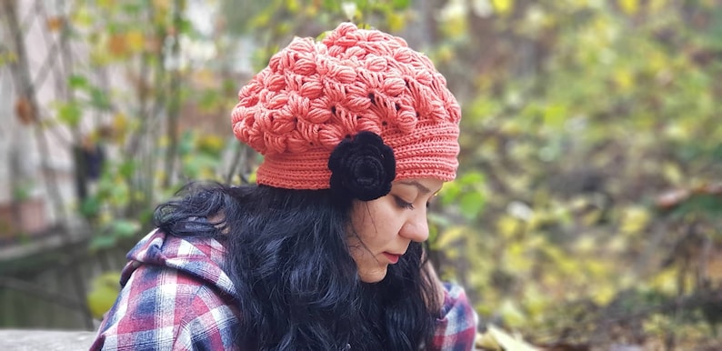 May include: A coral pink crocheted beanie with a black flower embellishment. The beanie is made with a textured stitch and has a slouchy fit.