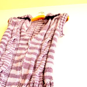 May include: A striped knitted vest with a cable knit pattern in shades of purple and white. The vest is hanging on a wooden hanger.