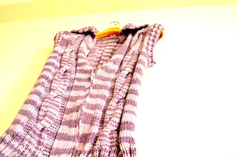 May include: A striped knitted vest with a cable knit pattern in shades of purple and white. The vest is hanging on a wooden hanger.