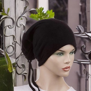 Op de afbeelding: Een zwarte fleece beanie met een lang, zwart trekkoord. De beanie is over het voorhoofd getrokken en heeft een losse pasvorm.