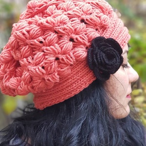 May include: A woman wearing a pink crocheted beanie with a black crocheted flower accent. She is wearing a red and black plaid shirt and has long black hair.