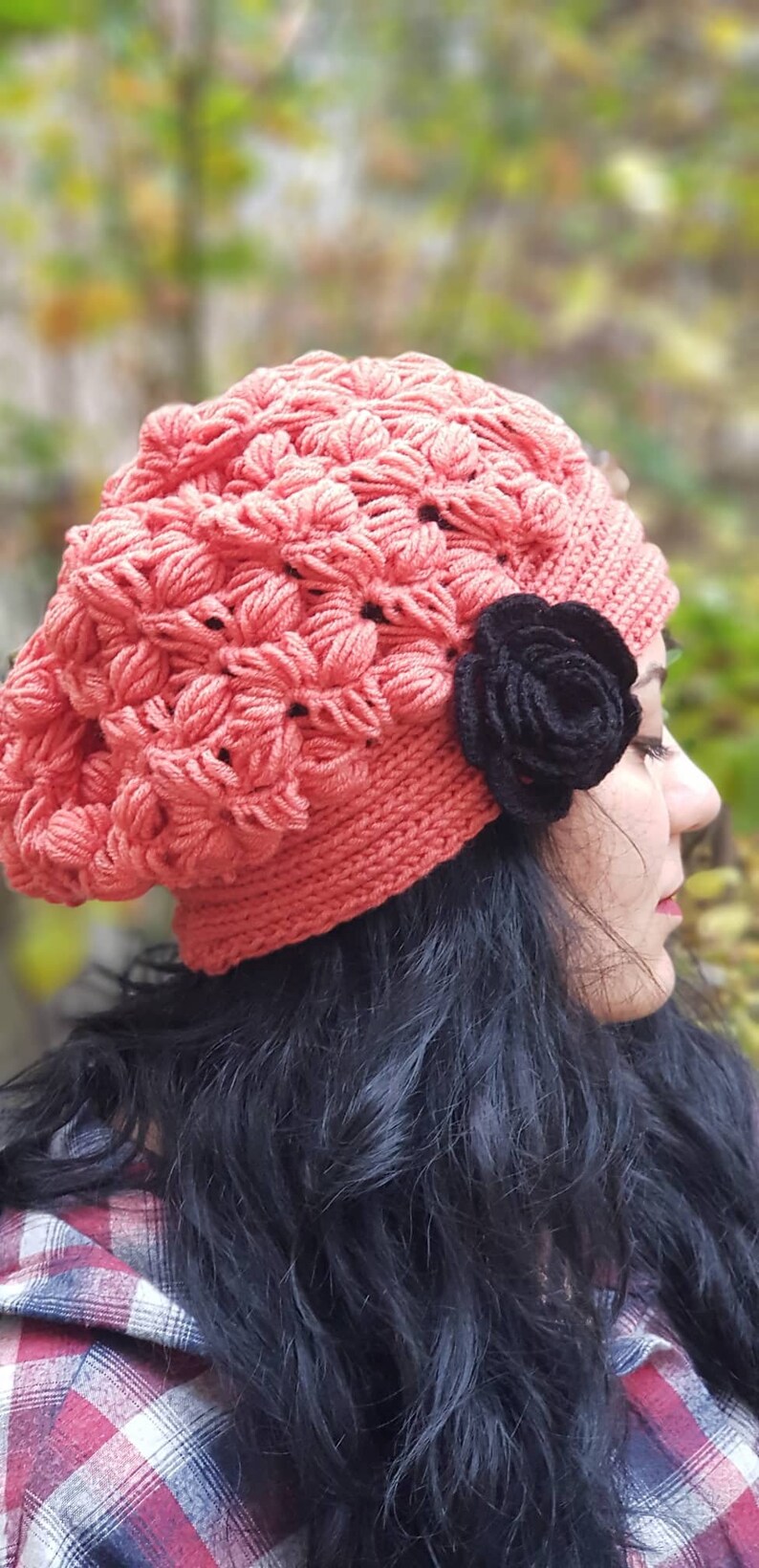 May include: A woman wearing a pink crocheted beanie with a black crocheted flower accent. She is wearing a red and black plaid shirt and has long black hair.