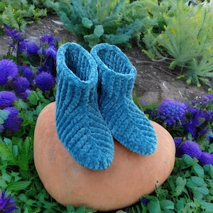 Op de afbeelding: Een paar tealblauwe gehaakte pantoffels met een textuurpatroon. De pantoffels liggen op een terracotta-kleurige steen.