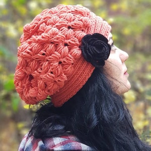 May include: A woman wearing a red crocheted beanie with a black flower accent. She is wearing a red and black plaid shirt and has long black hair.