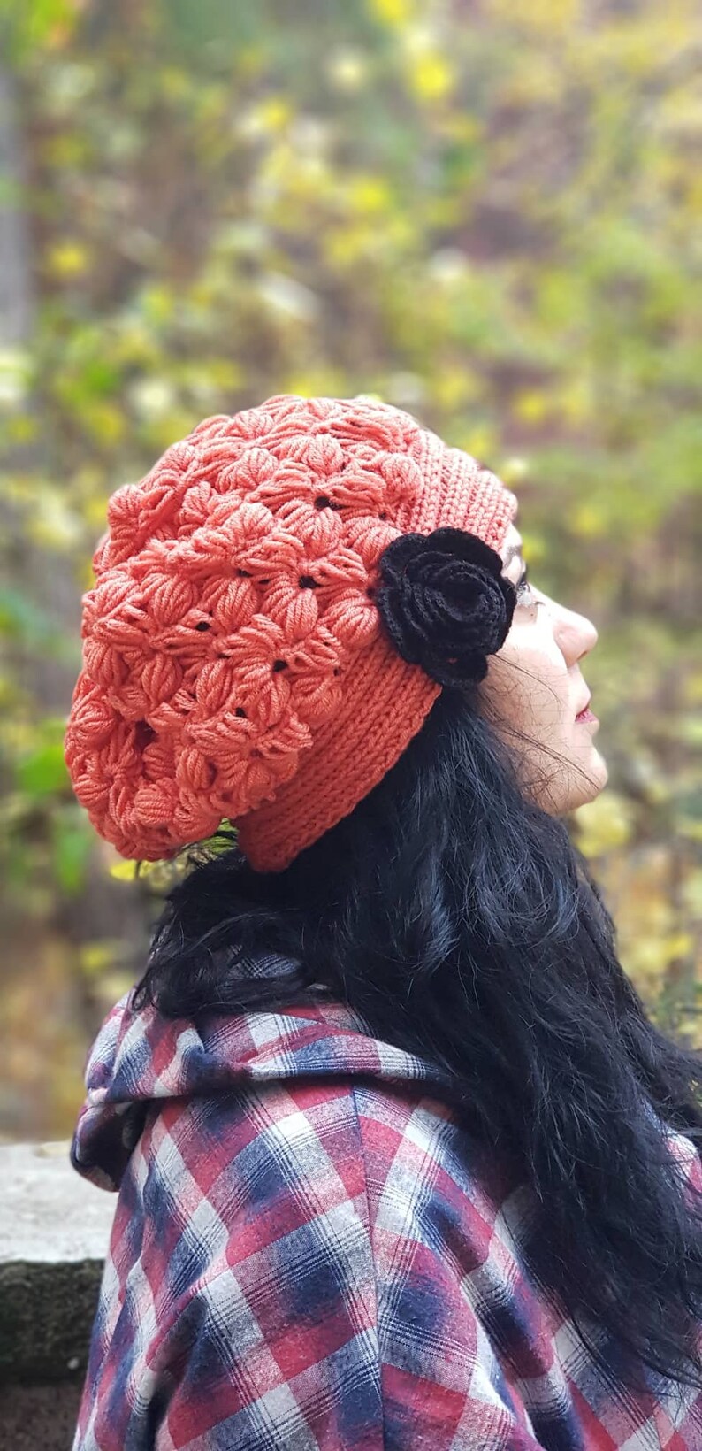 May include: A woman wearing a red crocheted beanie with a black flower accent. She is wearing a red and black plaid shirt and has long black hair.