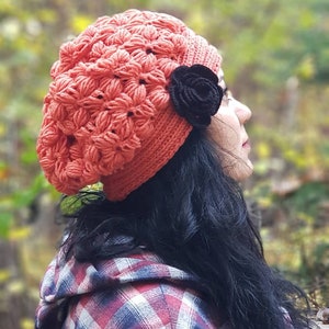 May include: A woman wearing a red crocheted beanie with a black flower accent. She is wearing a red and white plaid shirt.