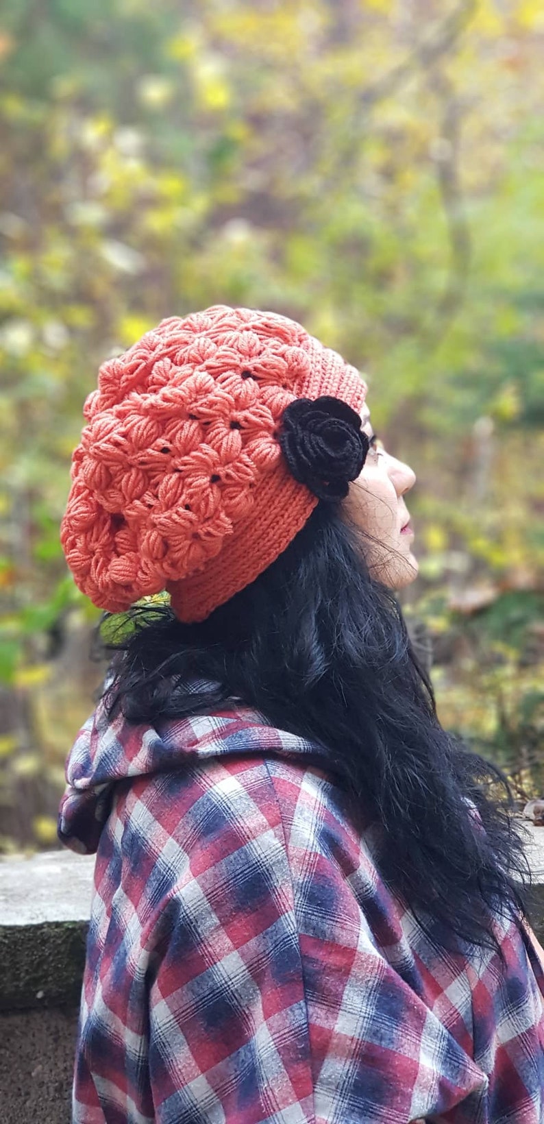 May include: A woman wearing a red crocheted beanie with a black flower accent. She is wearing a red and white plaid shirt.