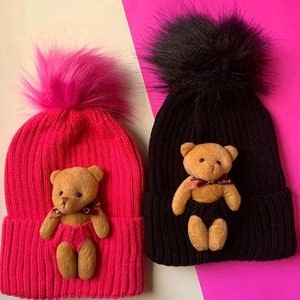 May include: Two knit winter hats with fuzzy pom poms. One hat is pink with a pink pom pom and a brown teddy bear applique. The other hat is black with a black pom pom and a brown teddy bear applique. Both teddy bears have a red ribbon around their necks that says "Love".