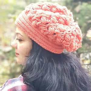 May include: A close-up of a woman wearing a coral crocheted beanie hat. The hat has a textured, puffy design. The woman has long, dark hair and is wearing a red and black plaid shirt.