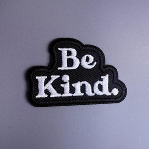 May include: A black embroidered patch with a cloud-like shape featuring the white text Be Kind. The patch has a thick black border and a textured fabric surface with visible stitching throughout the letters and the surrounding background area.