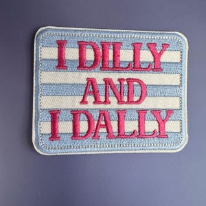 May include: A rectangular embroidered patch featuring horizontal light blue and white stripes. The text "I DILLY AND I DALLY" is stitched in bold, bright pink capital letters across the center of the patch, which has a finished white border.