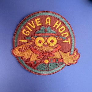 May include: A circular embroidered patch featuring a brown owl wearing a green ranger hat and uniform. The owl is saluting with one wing. The text I GIVE A HOOT is stitched in yellow letters along the top edge of the patch against a red background.