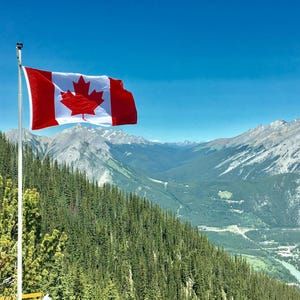 Könnte beinhalten: Eine kanadische Flagge mit rotem und weißem Design und einem Ahornblatt weht vor einer Bergkette und blauem Himmel. Im Vordergrund befindet sich ein mit grünen Bäumen bedeckter Hang.