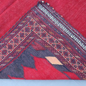 May include: A folded red rug with a detailed border. The border features geometric patterns in burgundy, cream, and teal. The rug's main field is a solid red, with a dark gray design in the corner.