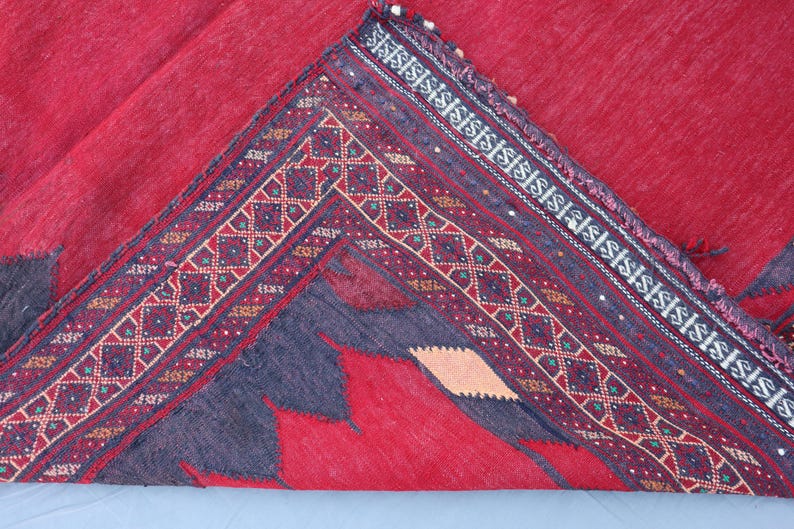 May include: A folded red rug with a detailed border. The border features geometric patterns in burgundy, cream, and teal. The rug's main field is a solid red, with a dark gray design in the corner.