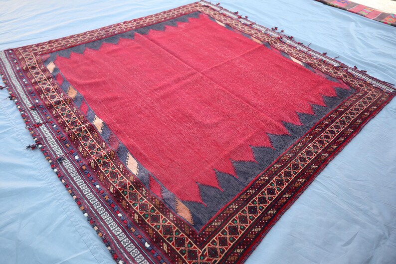 May include: A large, rectangular rug with a vibrant red center and a decorative border. The border features intricate geometric patterns in shades of brown, black, and cream. The rug has a dark blue zig-zag design around the red center.