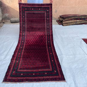 May include: A rectangular runner rug featuring a deep burgundy and black color palette. The central field displays a repeating geometric pattern, framed by a wide border with intricate linear motifs and small arrow-like shapes in dark tones.