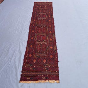 May include: A long, rectangular rug with a deep red background and intricate geometric patterns in shades of red, brown, and gold. The rug features a central design with repeating motifs and a fringed edge.