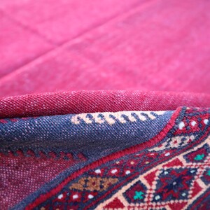 May include: Close-up of a section of a red and blue patterned rug. The rug's design features geometric shapes and floral motifs in red, blue, and cream colors. The rug is folded, revealing its layered construction.