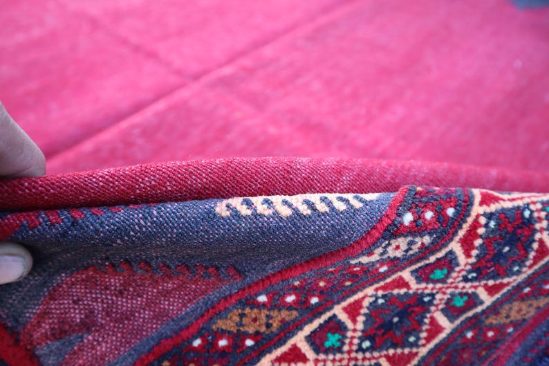 May include: Close-up of a section of a red and blue patterned rug. The rug's design features geometric shapes and floral motifs in red, blue, and cream colors. The rug is folded, revealing its layered construction.