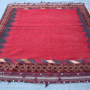 May include: A rectangular red rug with a dark blue and brown border. The rug has a central red field, a dark blue zig-zag pattern, and a detailed border with geometric designs and tassels. The rug is likely handmade.