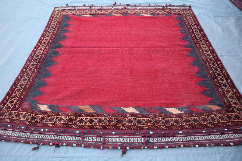 May include: A rectangular red rug with a dark blue and brown border. The rug has a central red field, a dark blue zig-zag pattern, and a detailed border with geometric designs and tassels. The rug is likely handmade.