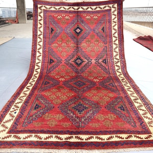 Pode incluir: Um grande tapete retangular vermelho com um padrão geométrico. O tapete apresenta formas de diamante e quadradas em tons de vermelho, castanho e azul. Uma borda creme com um design em zigue-zague emoldura o tapete. O tapete é exibido ao ar livre.