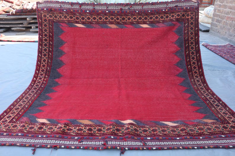May include: A large, rectangular rug with a vibrant red central field. The rug features a dark gray border with a zigzag pattern and intricate geometric designs in brown, gold, and other colors. Tassels are visible at the bottom.