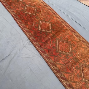 Puede incluir: Una alfombra larga y rectangular con un patrón geométrico de diamantes. La alfombra presenta una cálida paleta de colores de tonos rojos, naranjas y marrones. El diseño incluye formas de diamantes repetidas y detalles intrincados en el borde.