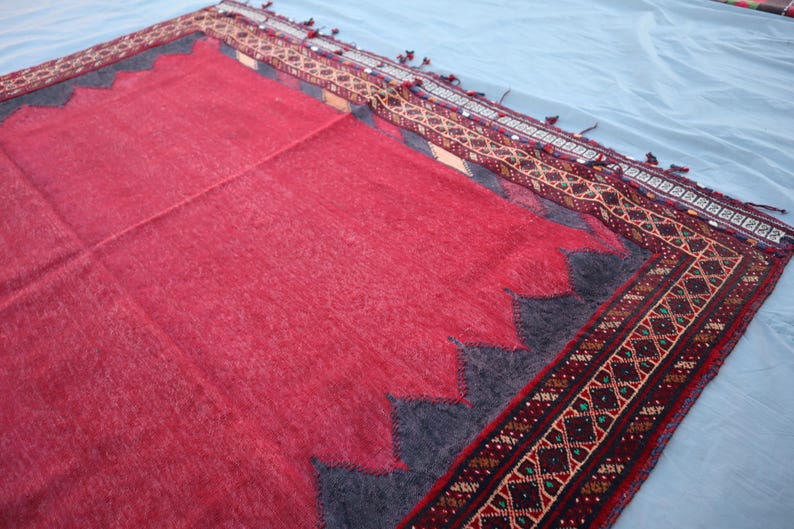 May include: A red rug with a dark gray zig-zag border and a patterned border in shades of red, brown, and green. The rug has a soft, textured appearance and is likely made of wool or a similar material. The rug is rectangular.