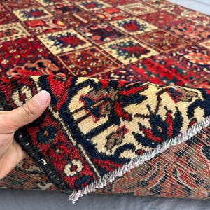 May include: A hand holds up a section of a patterned rug, revealing its intricate design. The rug features a repeating pattern of red, beige, and navy blue squares with floral motifs. The edges are finished with a dark border and fringe.