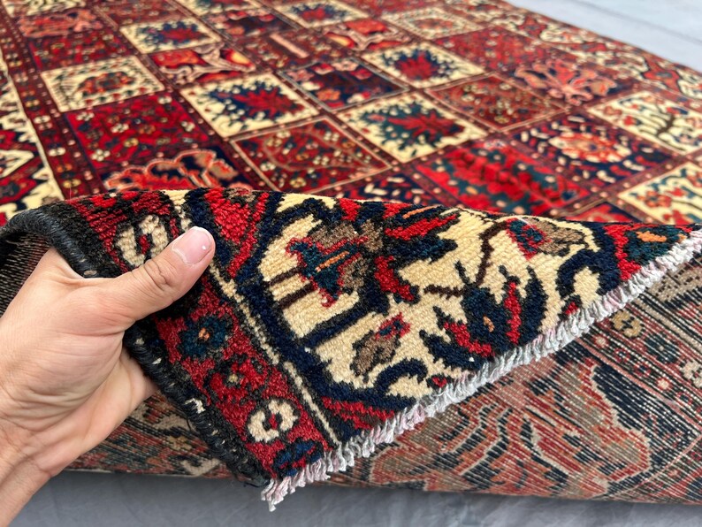 May include: A hand holds up a section of a patterned rug, revealing its intricate design. The rug features a repeating pattern of red, beige, and navy blue squares with floral motifs. The edges are finished with a dark border and fringe.
