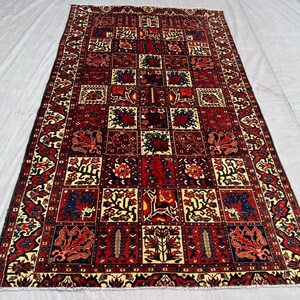 May include: Rectangular rug with a repeating geometric pattern in shades of red, cream, and navy blue. The design features squares filled with floral and abstract motifs, framed by a detailed border. The rug is likely made of wool.