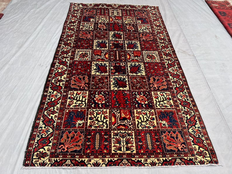 May include: Rectangular rug with a repeating geometric pattern in shades of red, cream, and navy blue. The design features squares filled with floral and abstract motifs, framed by a detailed border. The rug is likely made of wool.