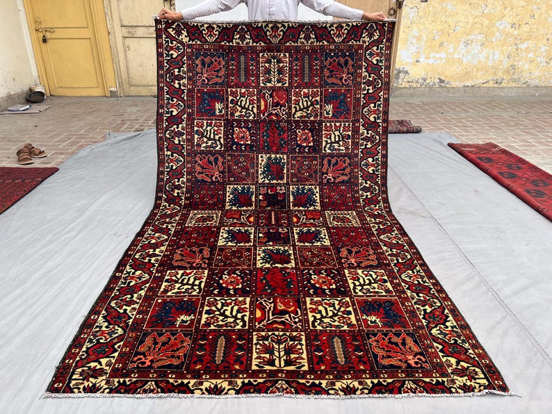 May include: A large, rectangular rug with a complex geometric pattern. The rug features a deep red background with repeating squares filled with floral and abstract designs in shades of blue, cream, and brown. The border has a detailed pattern.
