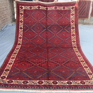 Pode incluir: Um tapete retangular grande com um fundo vermelho escuro e padrões geométricos intrincados. O design apresenta formas de diamante e uma borda com motivos florais e geométricos. O tapete tem uma franja creme ao longo da borda inferior.